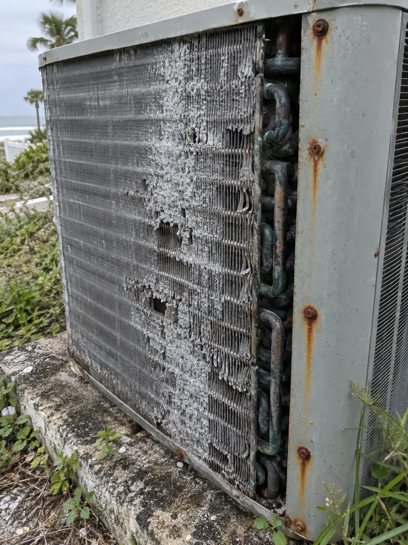 Corroded outdoor AC coil with crumbling white-coated aluminum fins, tarnished copper tube bends and rust streaks running from the cabinet screws